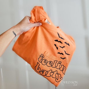 May include: Orange t-shirt with a pocket, featuring the phrase "feelin batty" in black script and a flock of black bats. The shirt is a solid orange colour.