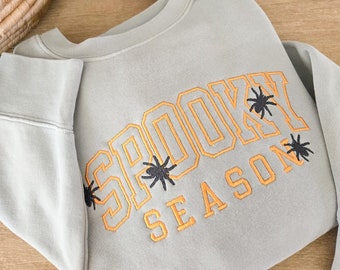 Puede incluir: Sudadera gris claro con las palabras "SPOOKY SEASON" bordadas en naranja con motivos de arañas negras. La sudadera está doblada sobre una superficie de madera, con una cesta de mimbre y una maceta gris oscuro en el fondo.