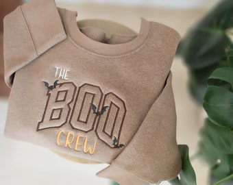 Puede incluir: Una sudadera de color beige con la frase bordada "THE BOO CREW" en marrón y amarillo, con motivos de murciélagos. La prenda doblada muestra el cuello redondo y las mangas largas. Un artículo de ropa con temática de Halloween.