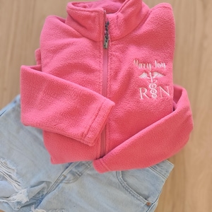May include: A vibrant pink fleece jacket with a full zip, embroidered with "Mary Joy R.N.", is neatly folded over light blue denim shorts with distressed details. The jacket is made of a soft, textured material, perfect for cooler days.