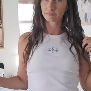 May include: A white tank top with a small embroidered design and blue denim shorts. The shorts have distressed details. The person is wearing white sneakers. The background includes framed photos and a bed with pillows.