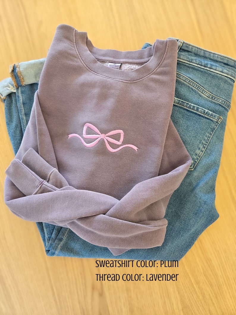 May include: Plum-colored sweatshirt with a lavender bow embroidery, paired with blue jeans. The sweatshirt's color is plum, and the thread color is lavender. The jeans are folded neatly next to the sweatshirt.