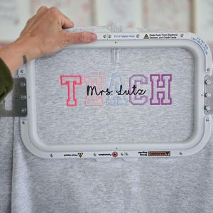 May include: A gray sweatshirt with the embroidered text "TEACH Mrs. Lutz" in pink, blue, and purple. The sweatshirt is held in a white embroidery hoop. The hoop has safety warnings printed on it.