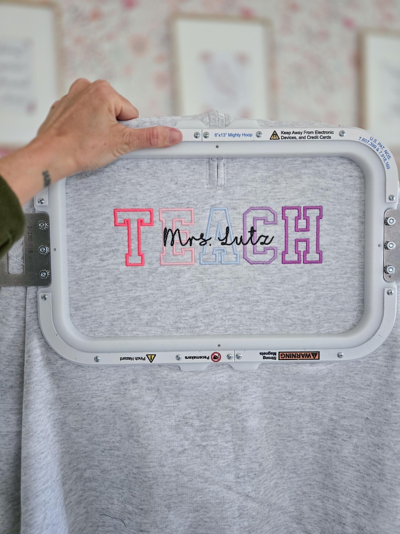 May include: A gray sweatshirt with the embroidered text "TEACH Mrs. Lutz" in pink, blue, and purple. The sweatshirt is held in a white embroidery hoop. The hoop has safety warnings printed on it.