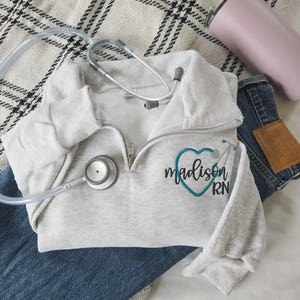 May include: A light grey quarter-zip sweatshirt with the name "Madison RN" embroidered in black and teal, a stethoscope, a pink tumbler, and blue jeans on a patterned blanket.