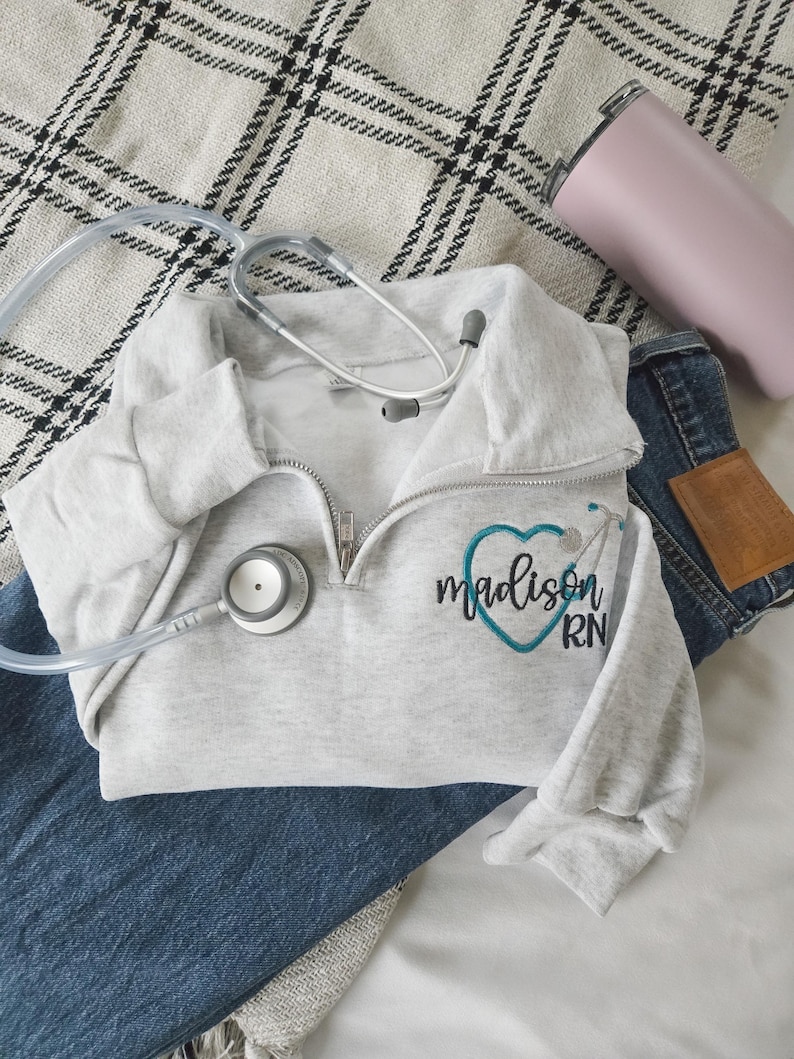 May include: A light grey quarter-zip sweatshirt with the name "Madison RN" embroidered in black and teal, a stethoscope, a pink tumbler, and blue jeans on a patterned blanket.