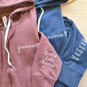 May include: Two zip-up hoodies, one in a dusty rose hue and the other in navy blue. The rose hoodie features "granna" embroidery, with names on the sleeve. The navy hoodie has "nonie" embroidered on the chest and names on the sleeve. Both have white drawstrings.