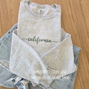 Könnte beinhalten: Ein hellgraues Sweatshirt mit dem Wort "california" in silbergrauem Salbeigarn gestickt. Das Sweatshirt ist über einer hellblauen Jeansshorts gefaltet. Das Sweatshirt ist aschfarben.