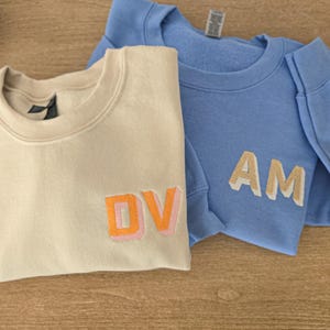 Op de afbeelding: Twee crewneck sweatshirts. Eén is crèmekleurig met de letters "DV" in oranje en roze. De andere is lichtblauw met "AM" in beige. Beide hebben een zachte, casual uitstraling.