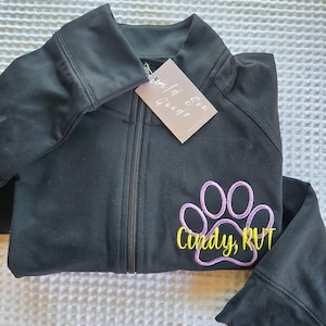 Op de afbeelding: Zwarte jas met ritssluiting en een pootafdrukontwerp in lichtpaarse en gele borduursels. De jas heeft een label met de tekst "Wild Sun Goods". Het pootafdrukontwerp bevat de naam "Cindy, RVT".