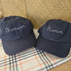 May include: Two navy blue baseball caps with white embroidered text. One cap says "Bronson" and the other says "Ronin". The caps are displayed on a patterned surface with a woven basket in the background.