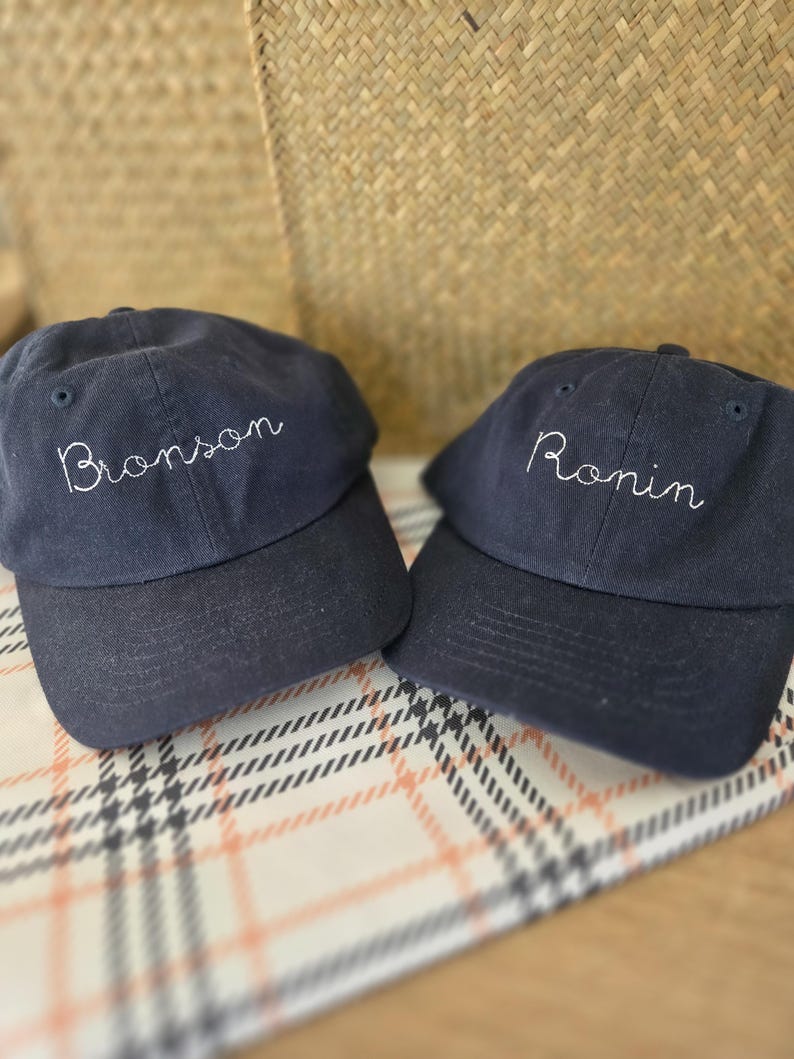 May include: Two navy blue baseball caps with white embroidered text. One cap says "Bronson" and the other says "Ronin". The caps are displayed on a patterned surface with a woven basket in the background.