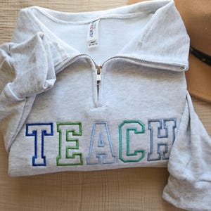 Puede incluir: Una sudadera gris claro con cuello con cremallera. La palabra "TEACH" est&aacute; bordada en el pecho con hilo azul y verde. La sudadera est&aacute; hecha de un material suave y tiene mangas largas.