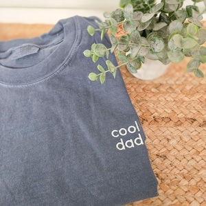 Peut inclure: Un t-shirt bleu ardoise à col rond avec une finition délavée. Le vêtement présente le texte "cool dad" brodé en fil blanc sur le côté inférieur droit de la poitrine. Le tissu présente une texture visible typique du coton teint en pièce.