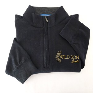 May include: A folded black quarter-zip sweatshirt with a gray collar. The sweatshirt has a gold embroidered design that reads "WILD SON Goods". The zipper is silver.