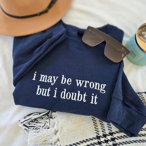 May include: Navy blue sweatshirt with the text "i may be wrong but i doubt it" in white. A brown fedora hat, sunglasses, and a candle are also in the image. The sweatshirt is folded on top of a patterned blanket.
