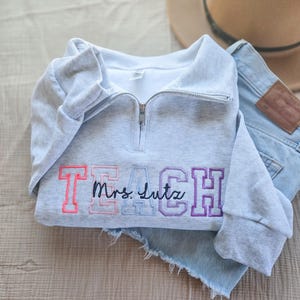 May include: A light gray sweatshirt with a quarter-zip neckline. The word "TEACH" is embroidered in pink, light blue, and purple, with "Mrs. Lutz" in cursive. A tan hat and denim shorts are in the background.