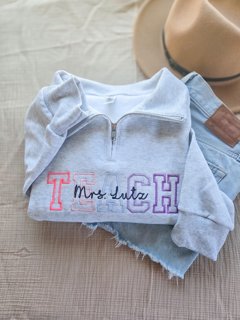 May include: A light gray sweatshirt with a quarter-zip neckline. The word "TEACH" is embroidered in pink, light blue, and purple, with "Mrs. Lutz" in cursive. A tan hat and denim shorts are in the background.