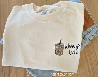 May include: Ivory t-shirt featuring an embroidered iced coffee design and the words "always late" in navy blue. The shirt is folded over denim shorts. The text "T SHIRT COLOR: IVORY" is at the bottom of the image.