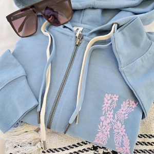 Puede incluir: Una sudadera con capucha azul claro con cremallera y un dise&ntilde;o floral "K" en rosa. La sudadera tiene una capucha con cord&oacute;n y una cremallera plateada. Gafas de sol oscuras descansan sobre la sudadera doblada.