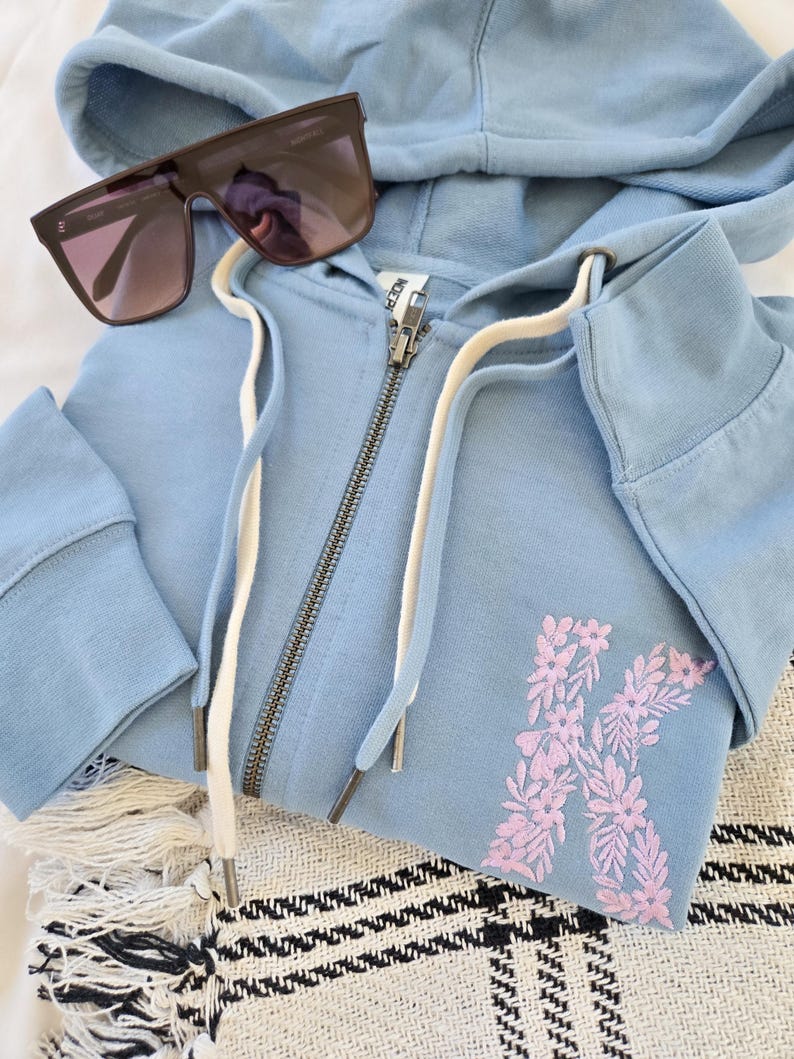 Puede incluir: Una sudadera con capucha azul claro con cremallera y un dise&ntilde;o floral "K" en rosa. La sudadera tiene una capucha con cord&oacute;n y una cremallera plateada. Gafas de sol oscuras descansan sobre la sudadera doblada.