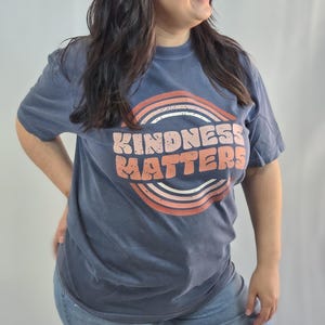 Puede incluir: Una camiseta azul marino con la frase "KINDNESS MATTERS" en un dise&ntilde;o retro. El texto es de color melocot&oacute;n, sobre un arco iris. La camiseta se combina con unos vaqueros azules.