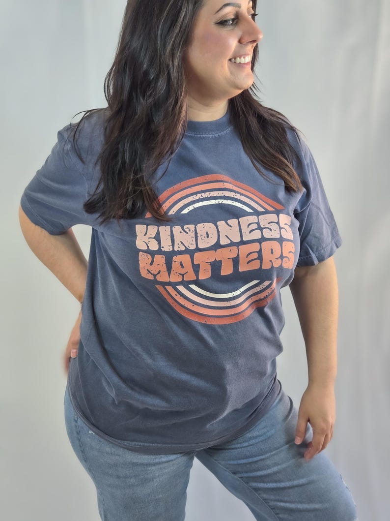 Puede incluir: Una camiseta azul marino con la frase "KINDNESS MATTERS" en un dise&ntilde;o retro. El texto es de color melocot&oacute;n, sobre un arco iris. La camiseta se combina con unos vaqueros azules.