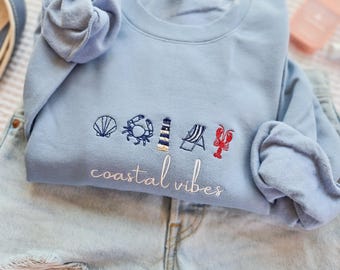 May include: A light blue sweatshirt with embroidered coastal themed designs, including a seashell, crab, lighthouse, beach chair, and lobster. The words "coastal vibes" are embroidered below the designs. The sweatshirt is folded on top of denim shorts.