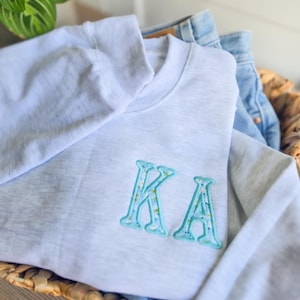 May include: A light gray sweatshirt with the embroidered letters "KA" in a light blue floral design. The sweatshirt is folded in a woven basket with a pair of blue jeans.