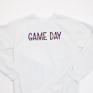 Pu&ograve; includere: Felpa bianca a maniche lunghe con la scritta "GAME DAY" in viola e giallo. La felpa ha un girocollo e maniche lunghe. Un capo casual per il giorno della partita.