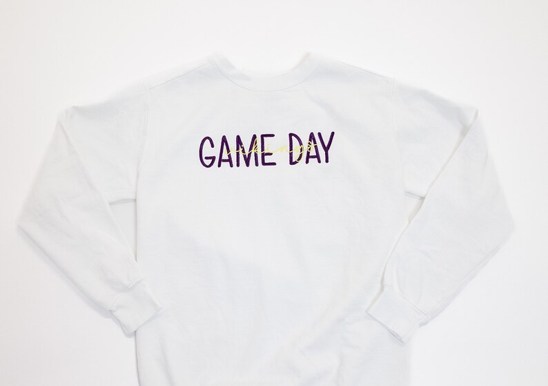 Pu&ograve; includere: Felpa bianca a maniche lunghe con la scritta "GAME DAY" in viola e giallo. La felpa ha un girocollo e maniche lunghe. Un capo casual per il giorno della partita.