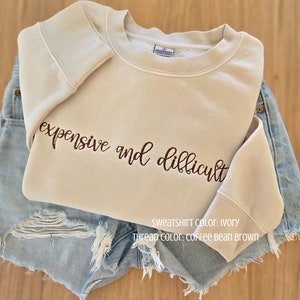 Könnte beinhalten: Elfenbeinfarbenes Sweatshirt mit der gestickten Aufschrift "expensive and difficult" in kaffeebraunem Garn. Das Sweatshirt liegt auf Jeansshorts. Das Bild enthält auch den Text "SWEATSHIRT COLOR: IVORY" und "THREAD COLOR: COFFEE BEAN BROWN".