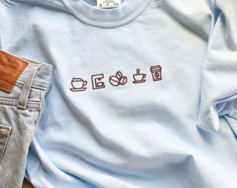 Puede incluir: Una camiseta Comfort Colors azul claro con un diseño bordado marrón con temática de café. El diseño incluye una taza de café, una cafetera, granos de café, una taza de café y una taza de café para llevar. Se ven unos vaqueros de mezclilla azul claro a la izquierda.