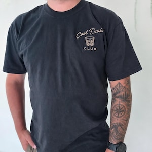 May include: A black short-sleeved t-shirt featuring a small graphic on the upper left chest. The graphic consists of a line drawing of a glass with ice cubes, positioned between the text "Cool Dads" above and "CLUB" below in a light tan color.