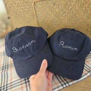 May include: Two navy blue baseball caps with white embroidered text. One cap says "Bronson" and the other says "Ronin". The caps are on a patterned surface.
