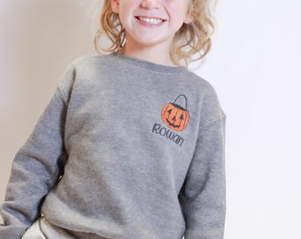 May include: A gray sweatshirt with a pumpkin design and the name "Rowan" embroidered on the front. The sweatshirt is paired with gray sweatpants. The pumpkin is orange with a black stem and face.