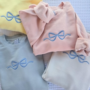 May include: Four pastel-colored sweatshirts are displayed, each featuring a light blue embroidered bow design. The colors include yellow, pink, blue, and off-white. The sweatshirts are made of a soft material and are neatly folded.