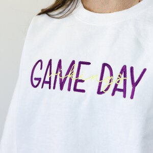 Pu&ograve; includere: Felpa bianca con la scritta ricamata "GAME DAY" in viola, con la parola "things" in corsivo giallo. Un capo d'abbigliamento casual.