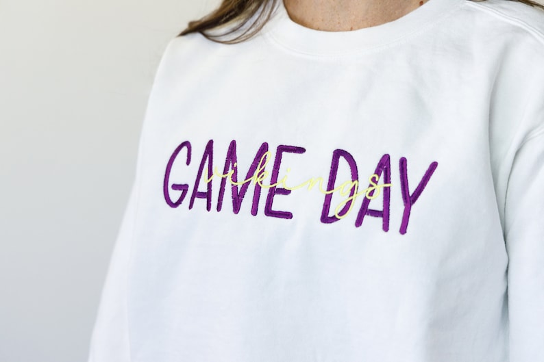 Pu&ograve; includere: Felpa bianca con la scritta ricamata "GAME DAY" in viola, con la parola "things" in corsivo giallo. Un capo d'abbigliamento casual.