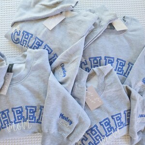 Könnte beinhalten: Mehrere graue Sweatshirts mit dem Wort "CHEER" in blauen Blockbuchstaben und "Mom" in weißer Schrift. Einige Ärmel sind mit Namen in blauer Schrift bestickt. An jedem Sweatshirt hängt ein Etikett.