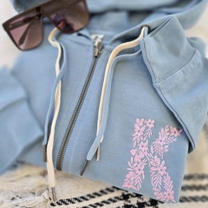 Puede incluir: Sudadera con capucha azul claro con cremallera y cord&oacute;n. Tiene un dise&ntilde;o floral rosa "K". Gafas de sol marrones descansan sobre la capucha. La sudadera est&aacute; doblada sobre una manta estampada en blanco y negro.
