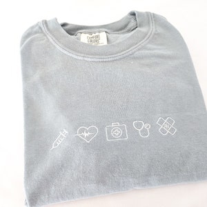 Puede incluir: Camiseta azul claro con s&iacute;mbolos m&eacute;dicos blancos bordados, incluyendo una jeringa, latido del coraz&oacute;n, botiqu&iacute;n, estetoscopio y tirita. La camiseta est&aacute; doblada.