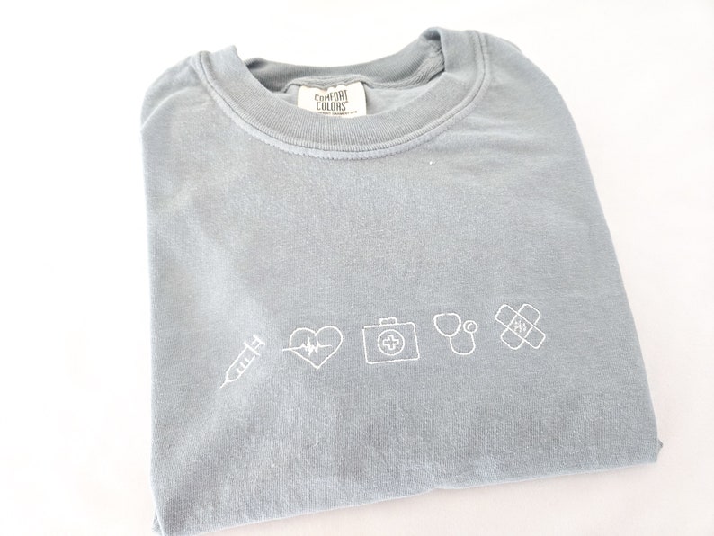 Puede incluir: Camiseta azul claro con s&iacute;mbolos m&eacute;dicos blancos bordados, incluyendo una jeringa, latido del coraz&oacute;n, botiqu&iacute;n, estetoscopio y tirita. La camiseta est&aacute; doblada.