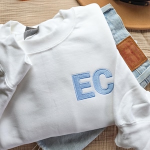 Può includere: Una felpa bianca con le lettere "EC" ricamate in azzurro. La felpa è piegata sopra un paio di pantaloncini di jeans. Un paio di occhiali con montatura arancione e un cappello marrone sono anch'essi nell'immagine.