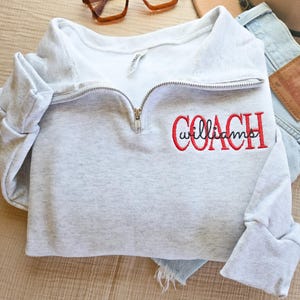 May include: A light gray sweatshirt with a quarter-zip neckline. The word "COACH" is embroidered in red, with "williams" below it. The sweatshirt is paired with brown-framed glasses and a hat.