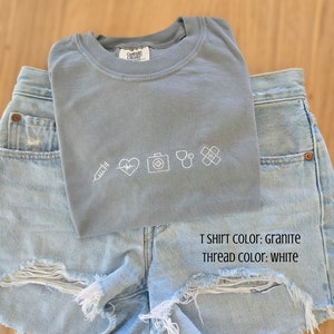 Puede incluir: Una camiseta gris Comfort Colors con s&iacute;mbolos m&eacute;dicos blancos bordados, incluyendo una jeringa, un coraz&oacute;n, un malet&iacute;n m&eacute;dico, un estetoscopio y una venda, se combina con shorts vaqueros azul claro. El texto en la camiseta dice "T SHIRT COLOR: GRANITE" y "THREAD COLOR: WHITE."
