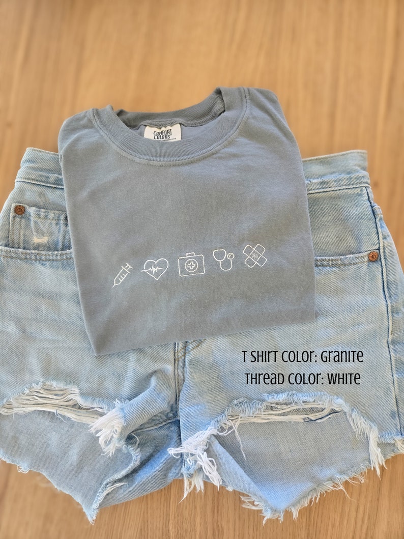 Puede incluir: Una camiseta gris Comfort Colors con s&iacute;mbolos m&eacute;dicos blancos bordados, incluyendo una jeringa, un coraz&oacute;n, un malet&iacute;n m&eacute;dico, un estetoscopio y una venda, se combina con shorts vaqueros azul claro. El texto en la camiseta dice "T SHIRT COLOR: GRANITE" y "THREAD COLOR: WHITE."