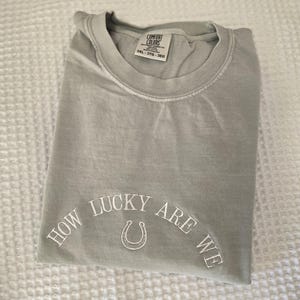Puede incluir: Una camiseta verde salvia doblada con el texto bordado "HOW LUCKY ARE WE" y un dise&ntilde;o de herradura. La camiseta es de la marca Comfort Colors, talla 3XL. La camiseta est&aacute; doblada sobre una superficie blanca texturizada.