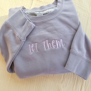 May include: A lavender-colored sweatshirt with the embroidered text "Let Them" in a cursive font. The sweatshirt has a crew neck and long sleeves with ribbed cuffs. The inside of the collar is a light gray color.