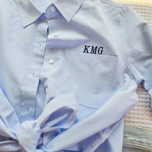 May include: Pale blue button-down shirt with a pocket and the embroidered initials "KMG" in navy blue. The shirt is tied at the waist, and part of a pair of blue jeans is visible.